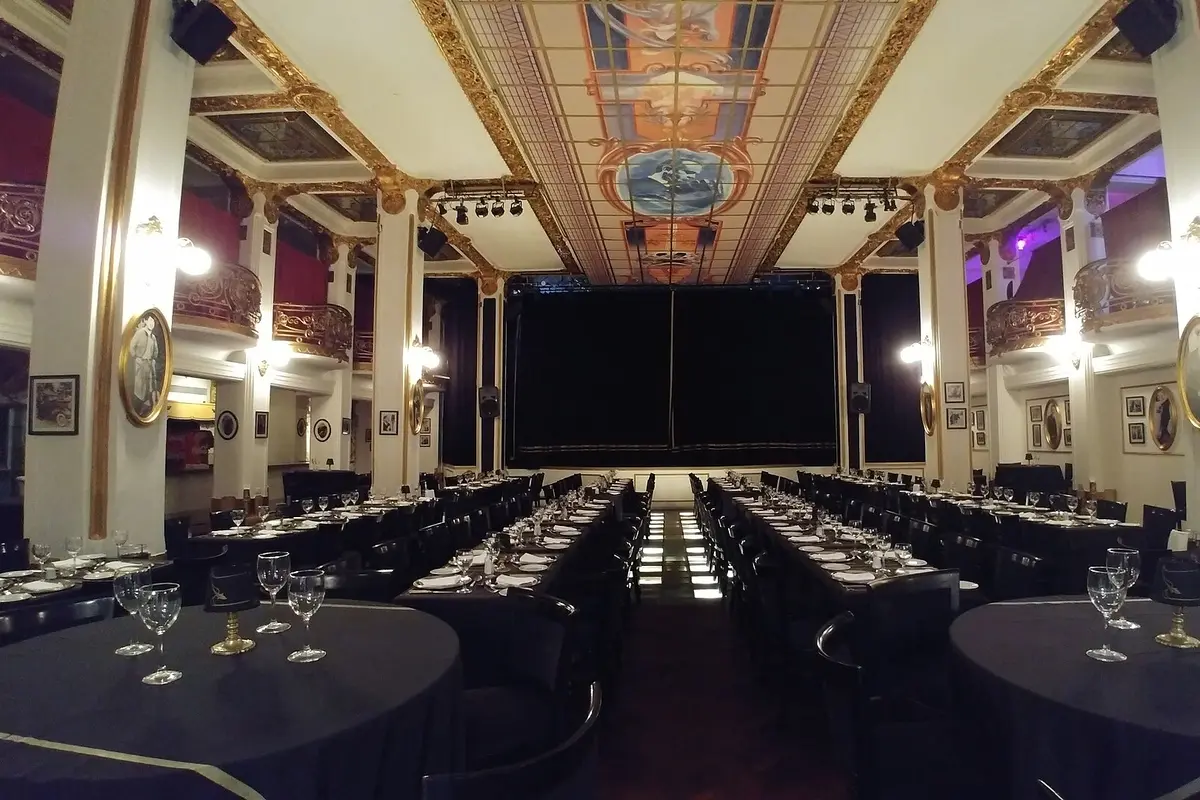 A tango room below the Galería Güemes laid with long dark tables and glasses facing a curtained stage, under a gilded and painted glass ceiling with private boxes along the sides